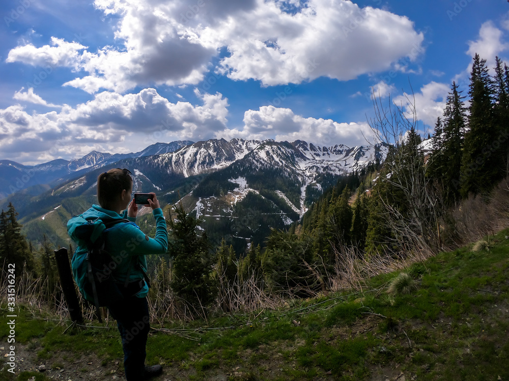 Woman with big hiking backpack taking a picture with a phone of the landscape, while hiking to Himmeleck peak, Austrian Alps. There is a massive mountain range in the back, partially covered with snow