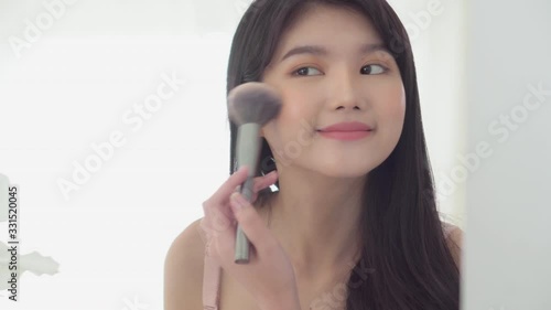 Beauty young asian woman happy and smile with face looking mirror applying makeup with brusher of cheek in the room, beautiful of girl holding brush make-up on facial, skin care and cosmetic concept. photo