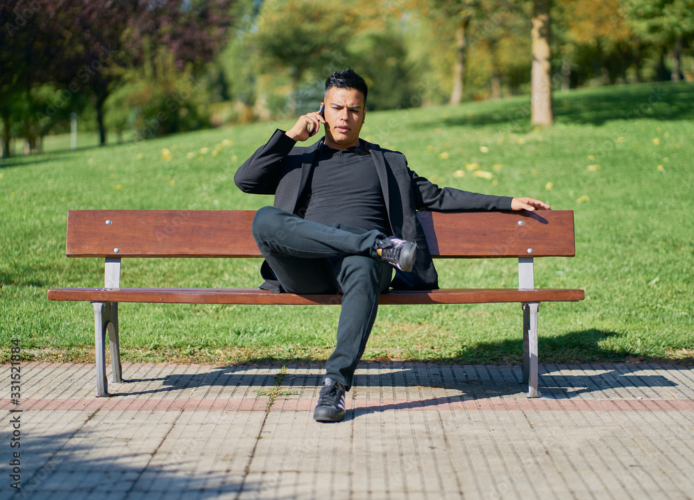 Hispanic man dressed as an executive in a black suit sitting on a park bench and talking on a cell phone