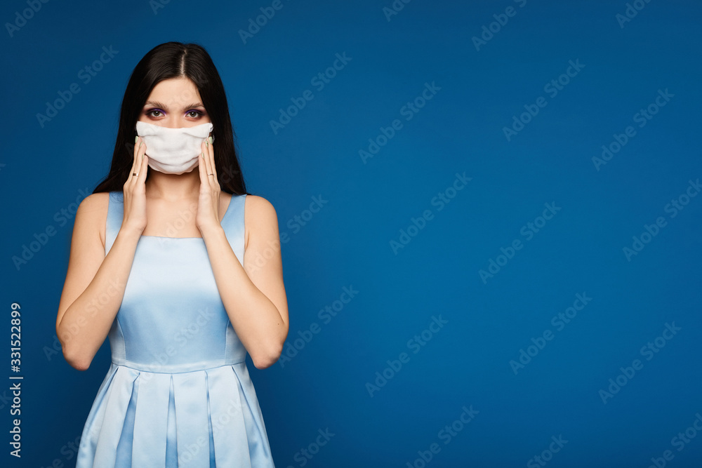 Young woman wearing a dress and medical mask, looking at the camera, isolated on a blue background. Brunette model girl in dress and face mask protective herself against flu epidemic, dust allergy.