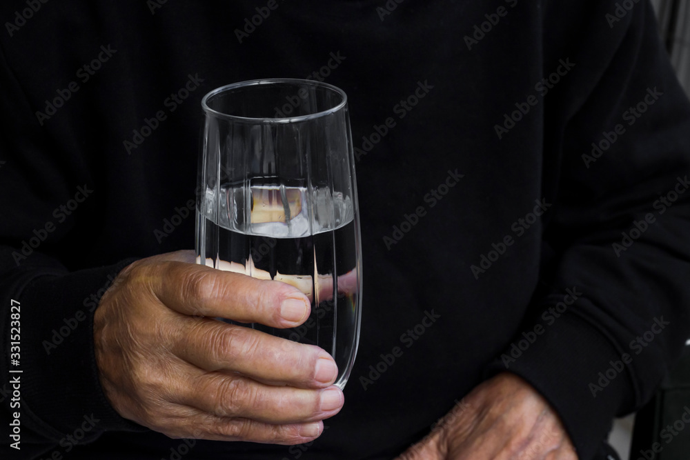 Hand of older person holding a glass of water on dark background.