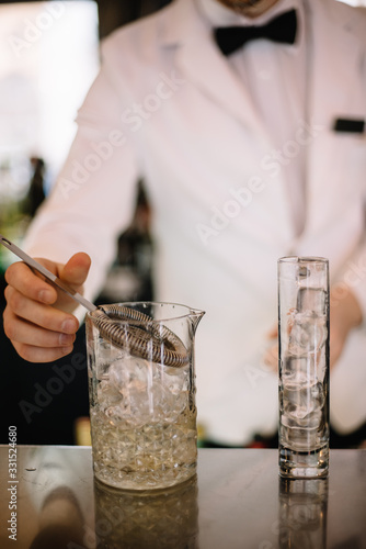 Bartender in a white suite and black bow tie preparing to pour a cocktail from a mixing pitcher into a highball glass with ice cubes using strainer. Smooth image with shallow depth of field.