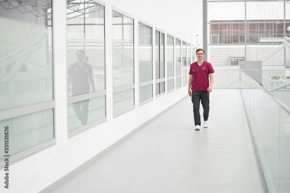 White smiling cheerful doctor wearing burgundy medical outfit going in corridors of clinic.