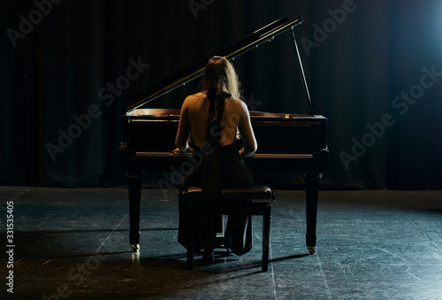 A woman from behind dressed in a black dress playing a black grand piano with the lid raised on a stage