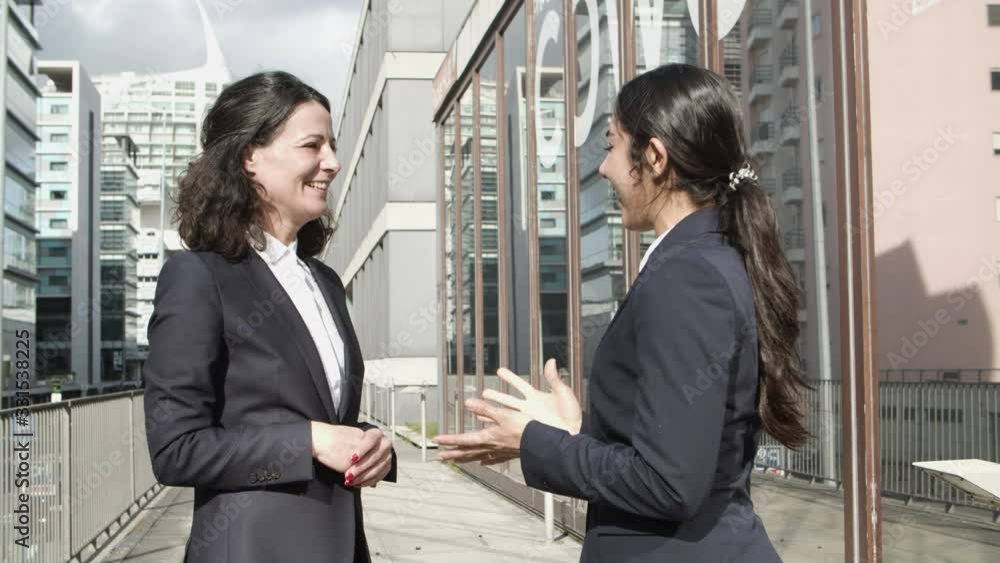 Businesswomen shaking hands and talking on street. Side view of cheerful multiethnic female colleagues shaking hands and talking outdoors. Business cooperation concept Businesswomen shaking hands and talking on street. Side view of cheerful multiethnic female colleagues shaking hands and talking outdoors. Business cooperation concept