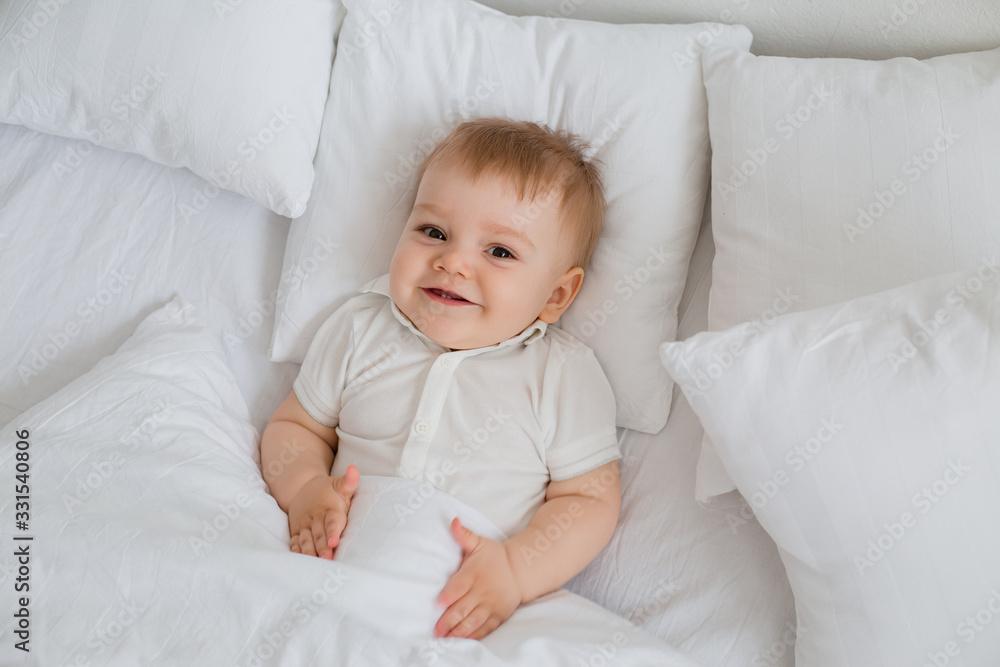Healthy toddler boy smiles in white bodysuit lies in bed with white linen