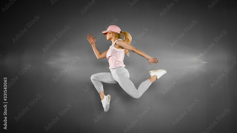 Young blonde woman with fit body jumping and running against grey background. Female model in sportswear exercising. Young blonde woman with fit body jumping and running against grey background. Female model in sportswear exercising.