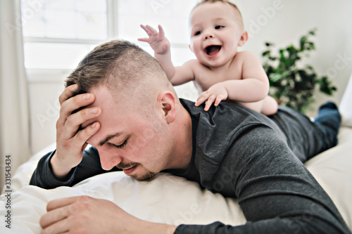 Tired father sleep but his baby boy interferes him. Young happy father lying on bed while his son play with him