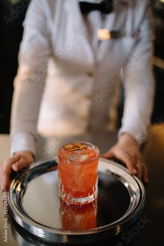 Female bartender holding on her hands metal tray with peach color cocktail in a rocks glass decorated with a dried slice of peach. Dark background. Smooth image with shallow depth of field.