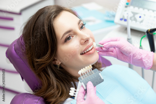  Doctor dentist checking and choosing teeth enamel shade for young woman.