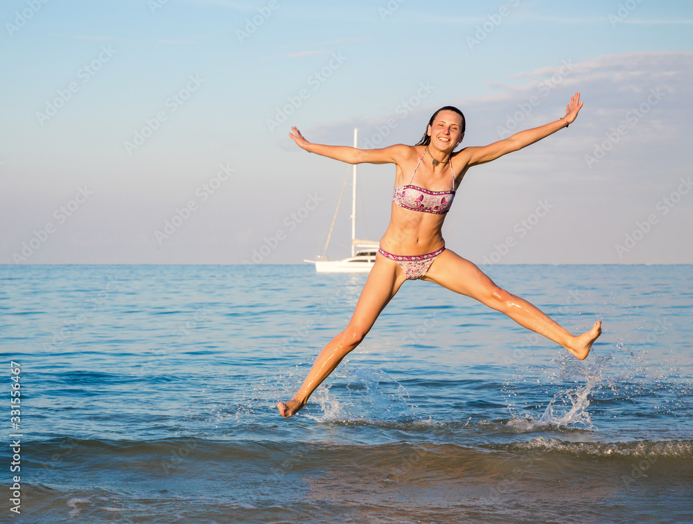 Happy young attractive woman in jump on the shore of beach at sunset day. White yacht in the distance in the background Happy young attractive woman in jump on the shore of beach at sunset day. White yacht in the distance in the background
