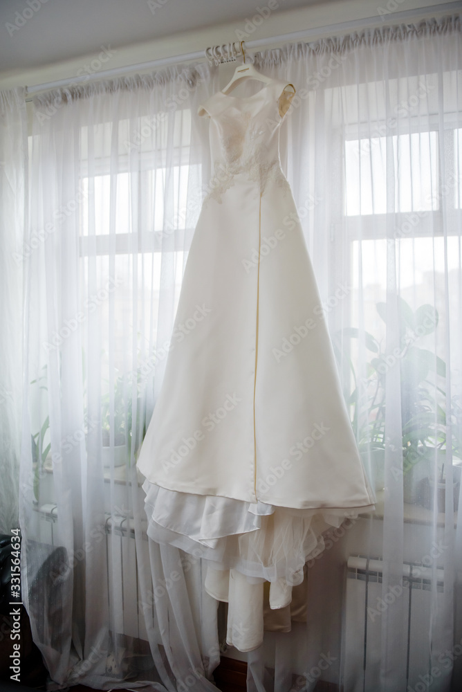 Elegant wedding white dress hanging on a window during a wedding preparation. Bride's morning. Before ceremony Elegant wedding white dress hanging on a window during a wedding preparation. Bride's morning. Before ceremony