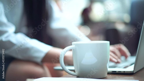 Ceramic coffee cup foreground and business female hands typing computer notebook keyboard background