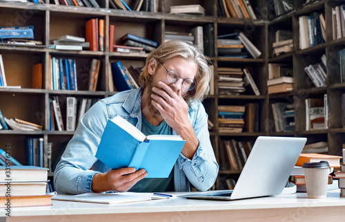 Obraz na plátně Bored tired sleepy guy university student yawning reading book studying in library feel lack of sleep of dull learning sit at desk preparing for college exam, fatigue of boring education concept