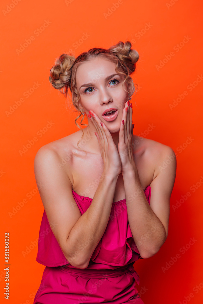 The girl in a red dress on a orange background in the studio. Blonde girl with two hair knots holding her chicks with hands