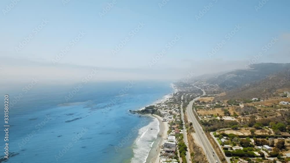 Drone camera shoots cloudy horizon, blue ocean, coastal settlement, winding road with cars and a mountain (Malibu, California, USA) Drone camera shoots cloudy horizon, blue ocean, coastal settlement, winding road with cars and a mountain (Malibu, California, USA)