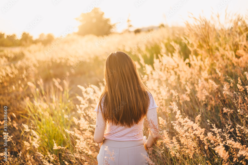 Back view of young woman walking in meadow grass field with warm sunshine. Conceptual of happiness woman enjoying her vacation.