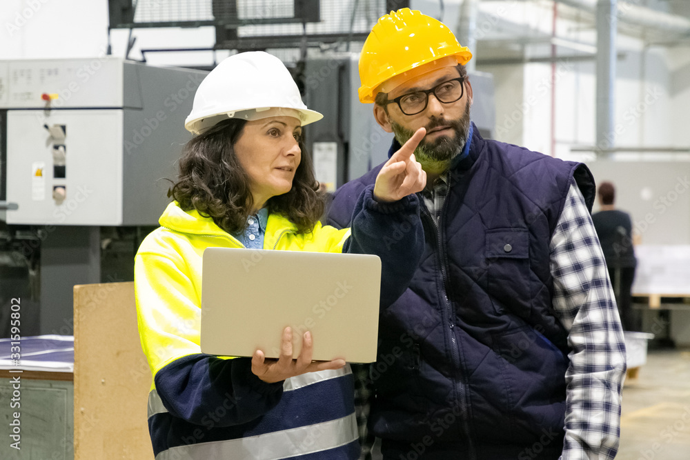 Two employees talking while standing with laptop at plant. Confident technicians discussing working process. Manufacturing concept Two employees talking while standing with laptop at plant. Confident technicians discussing working process. Manufacturing concept
