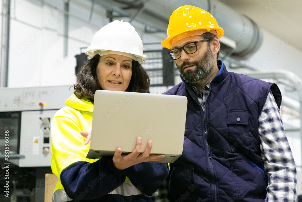 Front view of two employees talking while standing with laptop. Confident technicians discussing working process. Manufacturing concept Front view of two employees talking while standing with laptop. Confident technicians discussing working process. Manufacturing concept