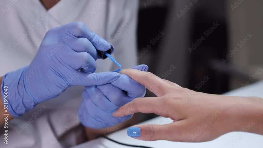 Master wears blue gloves. Nail artist puts blue nail polish using brush. Manicurist makes manicure for female client. Master wears blue gloves. Nail artist puts blue nail polish using brush. Manicurist makes manicure for female client.