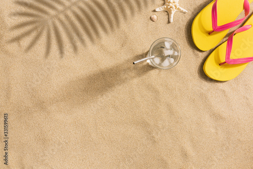 Summer fashion, summer outfit on sand background. Yellow flip flops and glass of water. Flat lay, top view. Harsh light with strong shadows