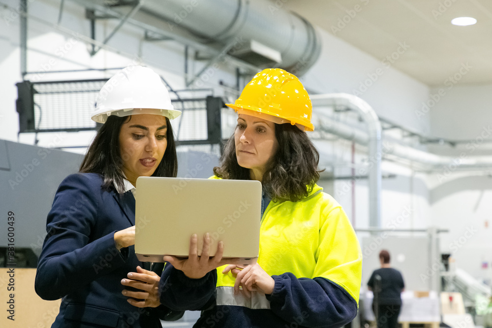 Front view of focused women looking at laptop at plant. Two female employees talking while standing at factory with laptop. Print manufacturing concept Front view of focused women looking at laptop at plant. Two female employees talking while standing at factory with laptop. Print manufacturing concept