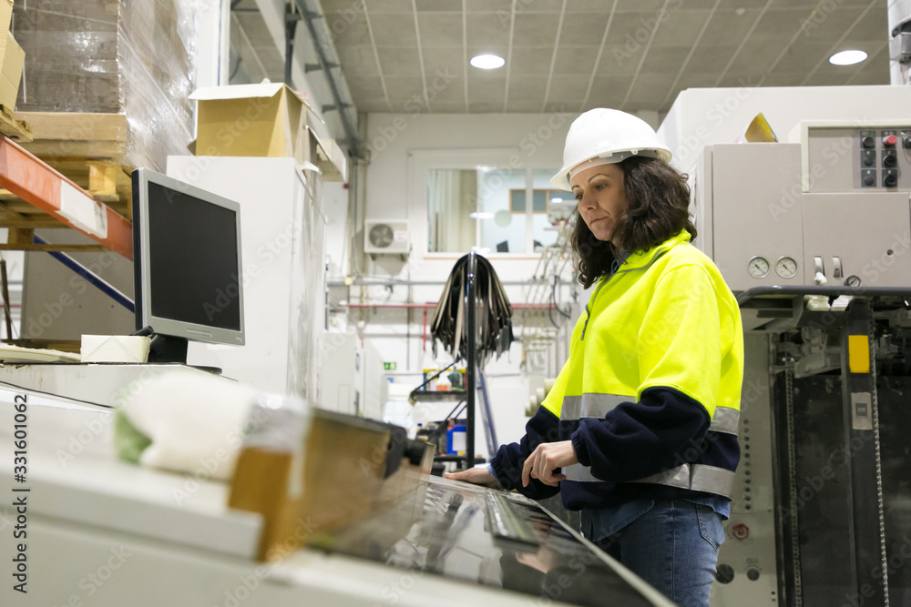 Focused female factory worker operating machine at control panel. Middle aged woman in uniform and helmet working at plant. Automated production concept Focused female factory worker operating machine at control panel. Middle aged woman in uniform and helmet working at plant. Automated production concept