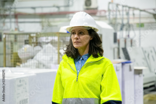 Concentrated mature woman in hard hat at manufacturing plant. Focused factory employee looking aside. Manufacturing concept