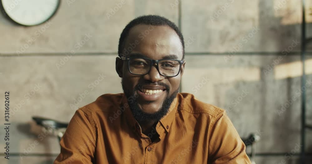 Portrait of cheerful African American office worker with glasses smiling to camera while sitting at table at workplace. Happy handsome office worker with smile on face indoors. Working concept Portrait of cheerful African American office worker with glasses smiling to camera while sitting at table at workplace. Happy handsome office worker with smile on face indoors. Working concept