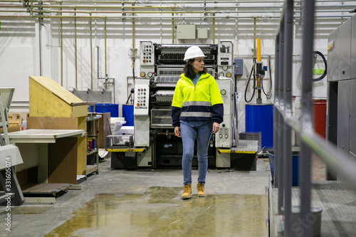 Confident female printing worker walking at factory. Mature woman in yellow hard hat walking near printing machine and looking aside. Print manufacturing concept