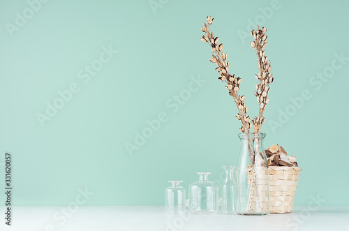 Contemporary interior in trendy mint menthe color with beige dry plant, flowers, wicker basket and glass vases on soft light white wood table.