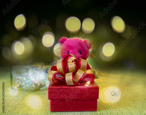 A red gift box with golden ribbon in a pink Teddy bear arms standing on green fabric,  a glass bottle in the back, under bokeh yellow lighting on dark background photo