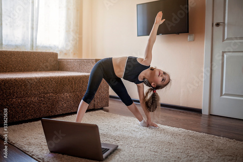 Pretty young girl in sportswear watching online video on laptop and doing fitness exercises at home photo