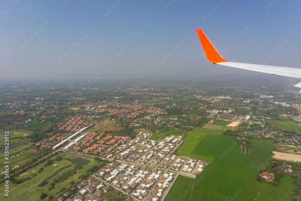 Sky scape view from clear glass window seat cabin crew to the aircraft wing of a plane, traveling on clouds and vivid blue sky above village city view and green farmer land