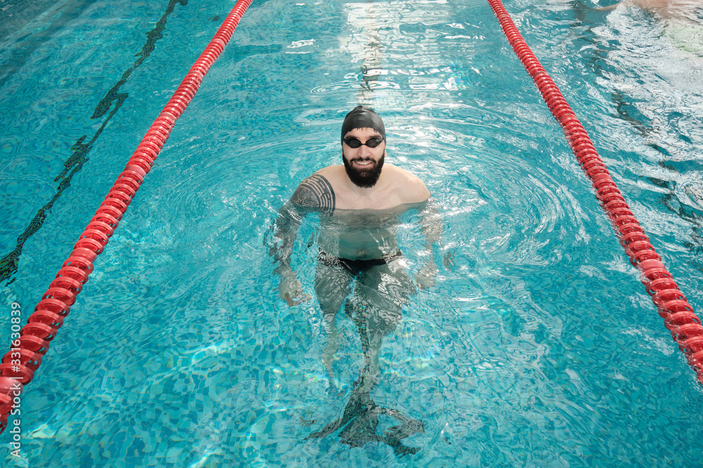Portrait of happy professional swimmer with beard wearing goggles and cap swimming in pool