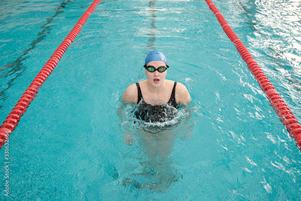 Portrait of young woman in swimming cap and goggles gliding out of water in pool