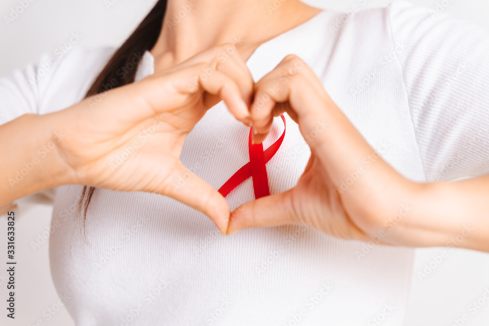 Closeup of woman hand in shape of heart with red badge ribbon on chest to support AIDS Day. Healthcare, medicine and AIDS awareness concept. Closeup of woman hand in shape of heart with red badge ribbon on chest to support AIDS Day. Healthcare, medicine and AIDS awareness concept.