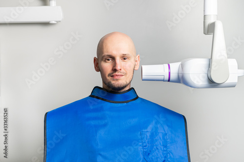 dental x-ray room. Portrait of a young bald man in a protective lead apron next to an X-ray machine during diagnosis.