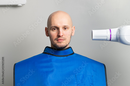 young bald man in a blue protective lead apron in an x-ray room near an x-ray apparatus for dental imaging. dentistry