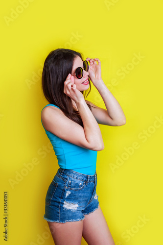 Cute young woman in blue T-shirt , denim shorts and sunglasses posing over yellow background.