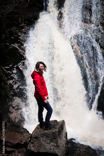 Junge Frau steht mit geschlossenen Augen und wehenden Haaren vor einem Wasserfall