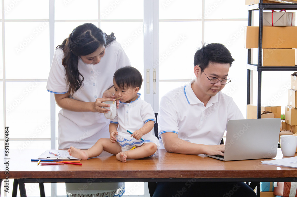 Asian young family together feeding adorable cute girl at home, father making small business marketing online logistic home office, mother playing with the child, parenthood duty multi task concept Asian young family together feeding adorable cute girl at home, father making small business marketing online logistic home office, mother playing with the child, parenthood duty multi task concept
