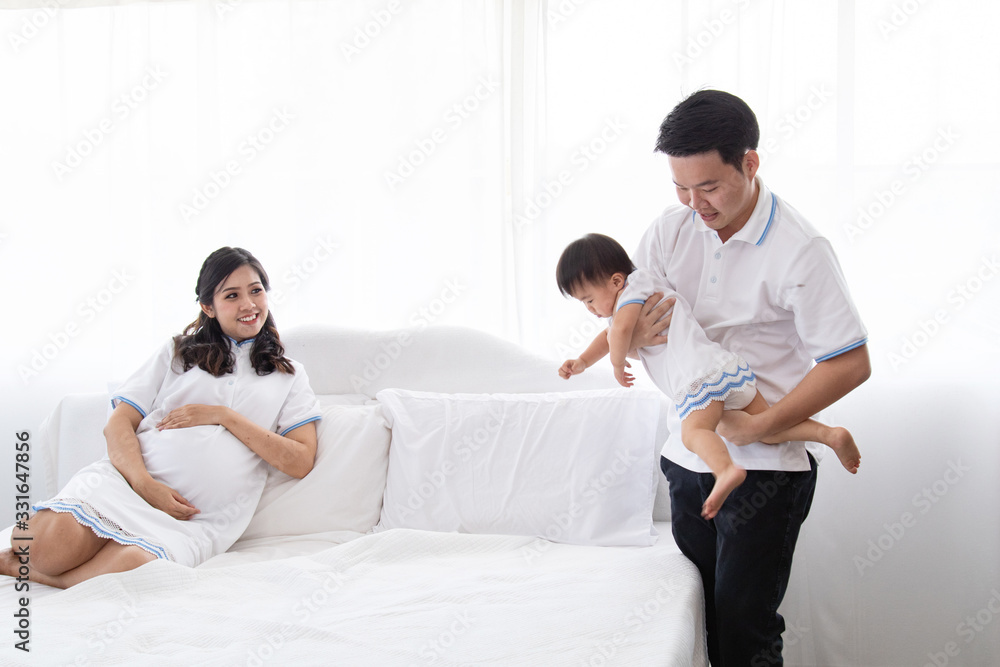 Asian young family help together feeding adorable cute girl at home, father playing with daughter hold her to fly on bed, mother pregnancy looking beloved husband and daughter multi task concept Asian young family help together feeding adorable cute girl at home, father playing with daughter hold her to fly on bed, mother pregnancy looking beloved husband and daughter multi task concept