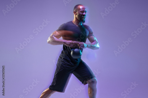 Emotional. Young african-american bodybuilder training over purple background in neon, mixed light. Muscular model with weight. Concept of sport, bodybuilding, healthy lifestyle, motion and action.