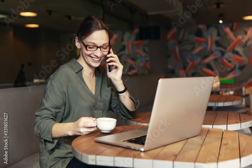 Attractive businesswoman in black jacket and stylish glasses sitting in cafe, talking on the phone and looking at laptop.