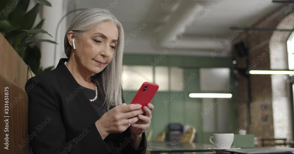 Side view of old businesswoman in earpods using smartphone and typing email. Modern mature elegant lady sitting in cafe and writing message. Concept of people, communication and tech. Side view of old businesswoman in earpods using smartphone and typing email. Modern mature elegant lady sitting in cafe and writing message. Concept of people, communication and tech.