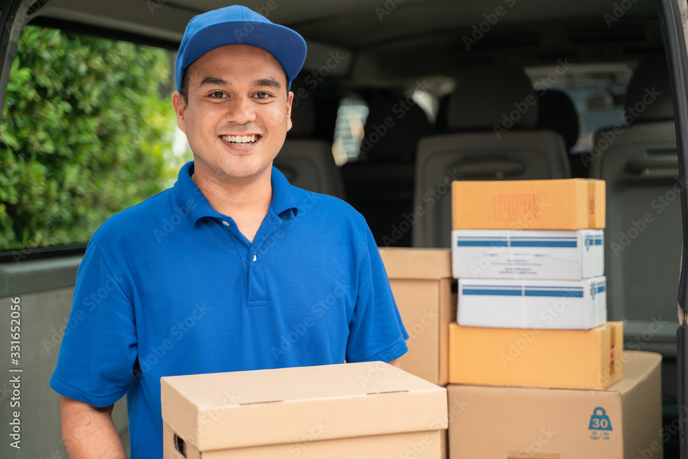 A smiling young asian delivery in blue uniform with parcel cardboard in front of customer house. Messenger and delivery concept. A smiling young asian delivery in blue uniform with parcel cardboard in front of customer house. Messenger and delivery concept.