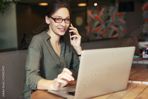 Attractive businesswoman in black jacket and stylish glasses sitting in cafe, talking on the phone and looking at laptop.