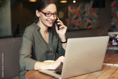 Attractive businesswoman in black jacket and stylish glasses sitting in cafe, talking on the phone and looking at laptop.