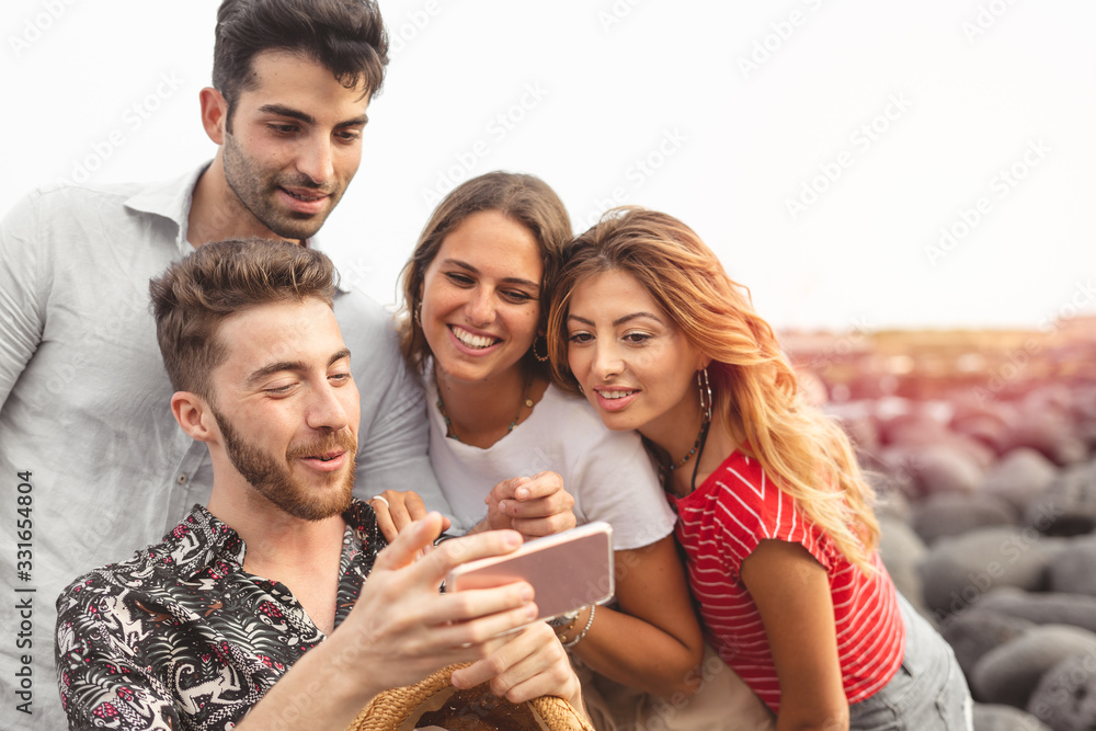 Obraz premium Young friends sitting outdoors and looking at smartphone. Young man showing something interesting to friends outdoors at the seaside